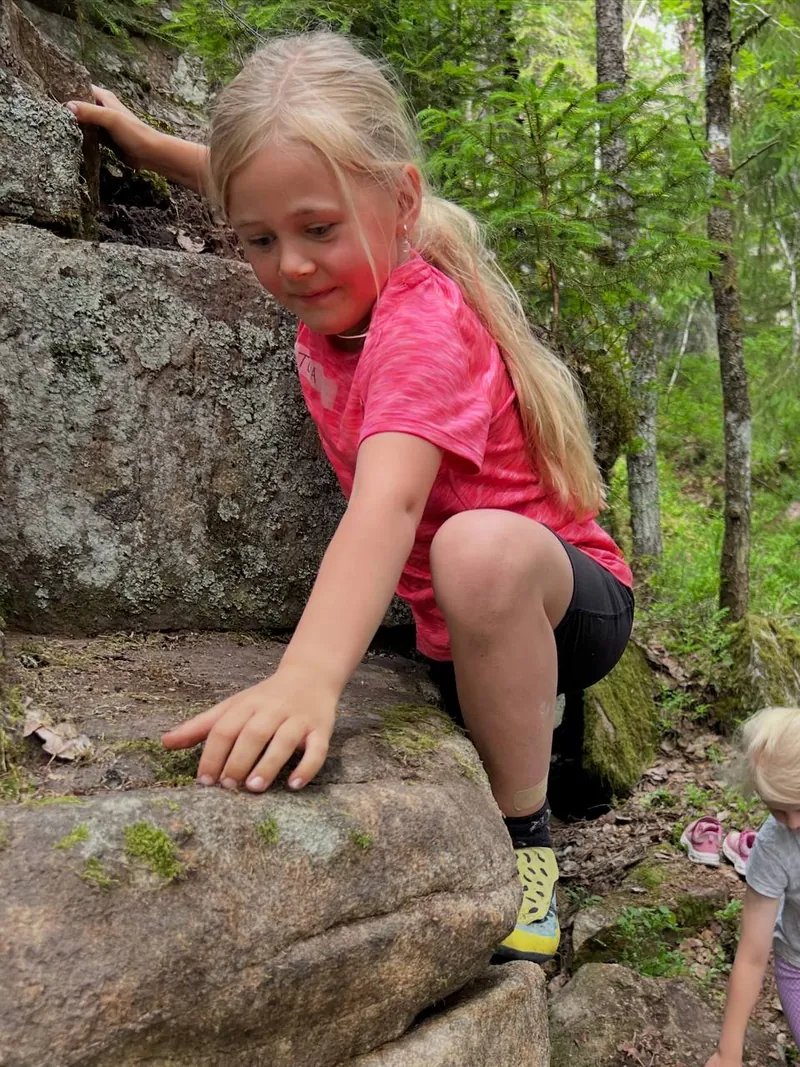 Barn i rosa skjorte klatrer på en stor stein mellom trær